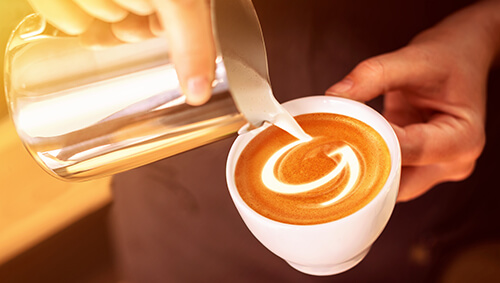 Person gießt Milch aus einer Milchkanne in eine Cappuccinotasse. Es entsteht Latte Art mit einem "G"