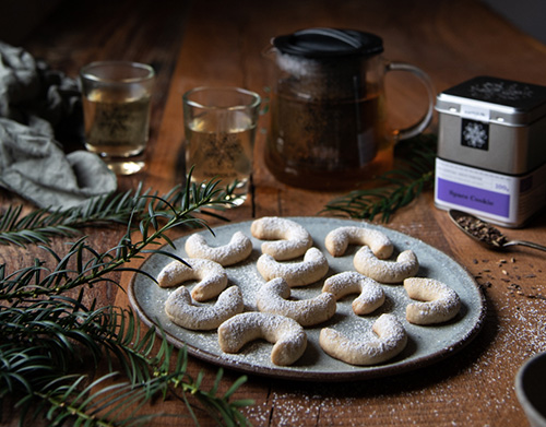 Weihnachtlicher Tisch mit einer Blechdose samova Space Cookie-Tee, gefüllter Teekanne, Teegläsern, Tannenzweigen und einem Blech voll Space-Cookie-Chai-Vanillekipferl