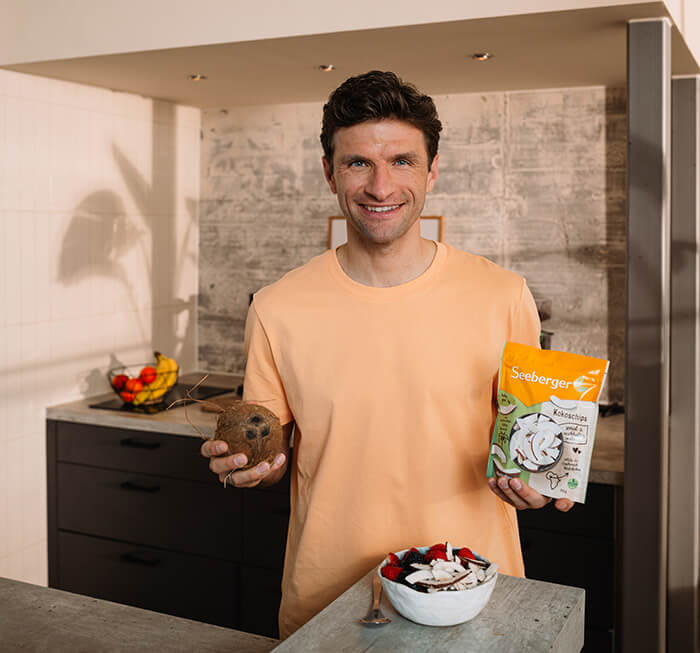 Thomas Müller hält eine Kokosnuss und eine Packung Seeberger Kokoschips